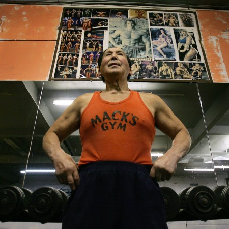 A muscular older man in an orange "Mack's Gym" tank top stands in a gym, with bodybuilding posters above him.