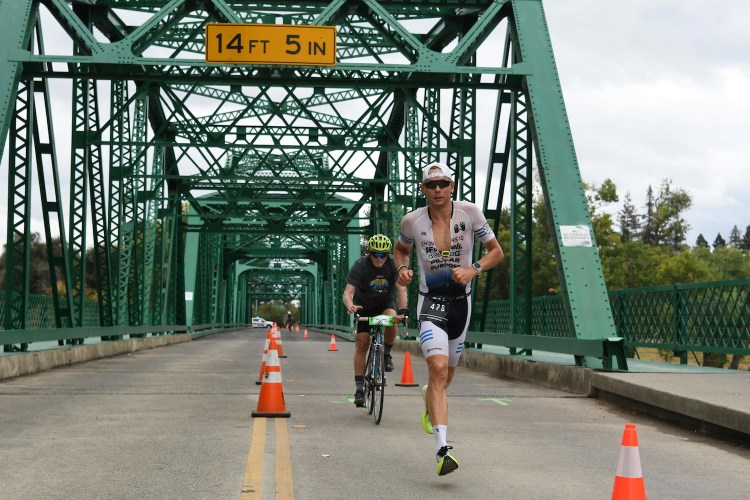 A triathlete in a white and black suit runs on a bridge, followed by a cyclist, with orange cones lining the road.