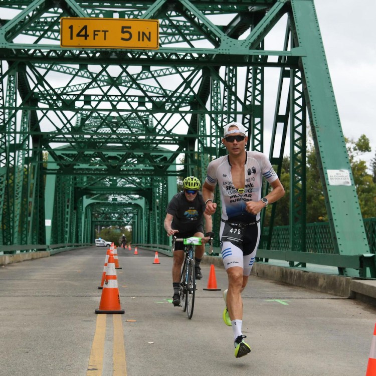 A triathlete in a white and black suit runs on a bridge, followed by a cyclist, with orange cones lining the road.