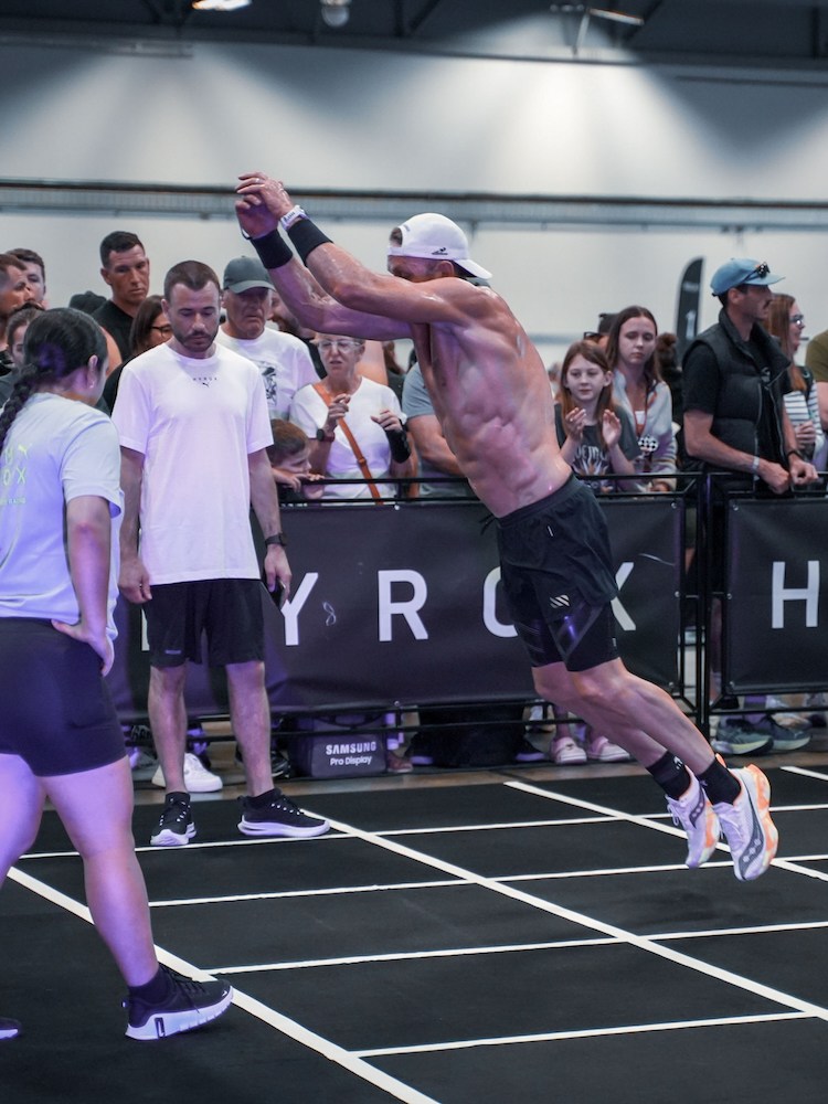 A shirtless man in a white cap and black shorts leaps horizontally, arms raised, on a black mat with white lines.