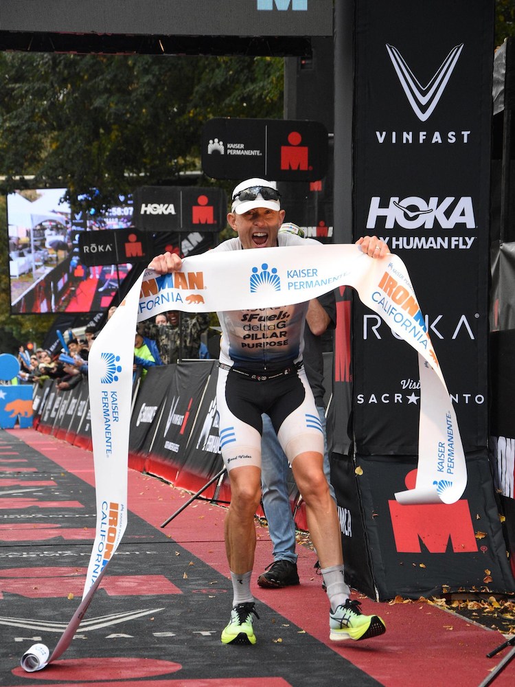 Ecstatic male triathlete in white and black gear, holding a finish line banner, at the Ironman California race.