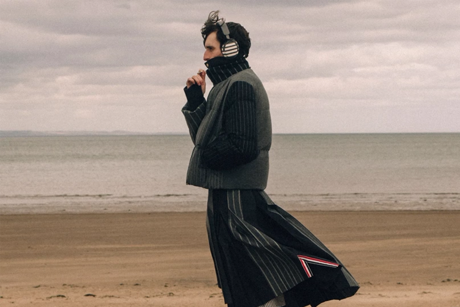 Man in a striped puffer vest, turtleneck, and pleated skirt with headphones on a sandy beach.
