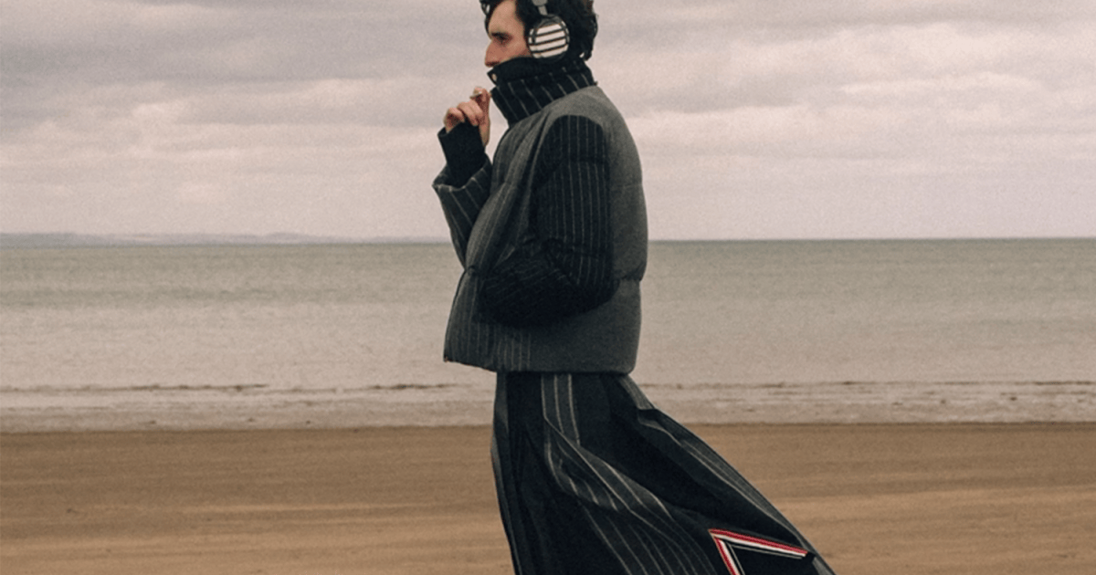 Man in a striped puffer vest, turtleneck, and pleated skirt with headphones on a sandy beach.