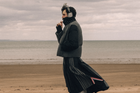 Man in a striped puffer vest, turtleneck, and pleated skirt with headphones on a sandy beach.