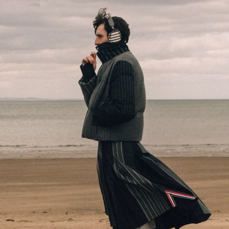 Man in a striped puffer vest, turtleneck, and pleated skirt with headphones on a sandy beach.