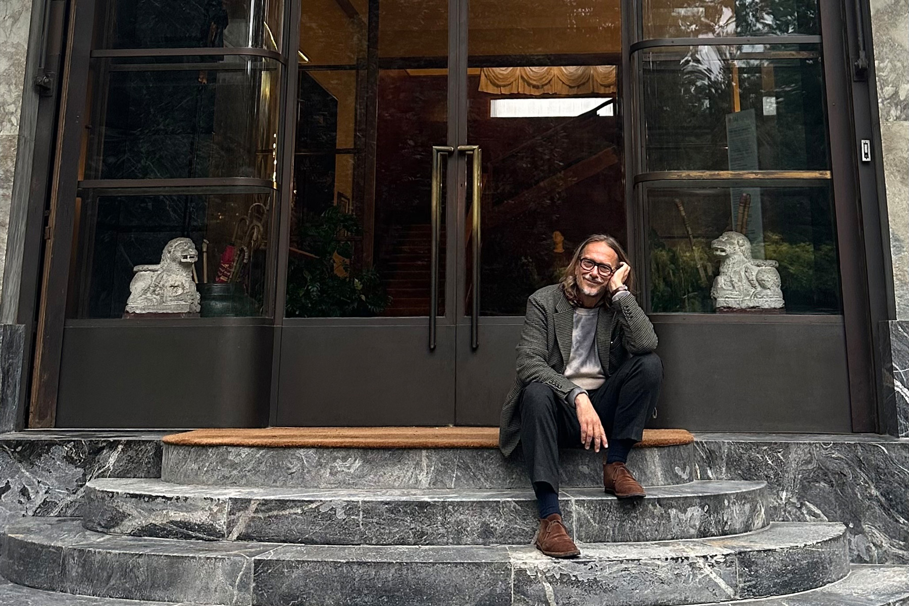 A smiling man with glasses and long hair sits on marble steps in front of a glass door with lion statues.