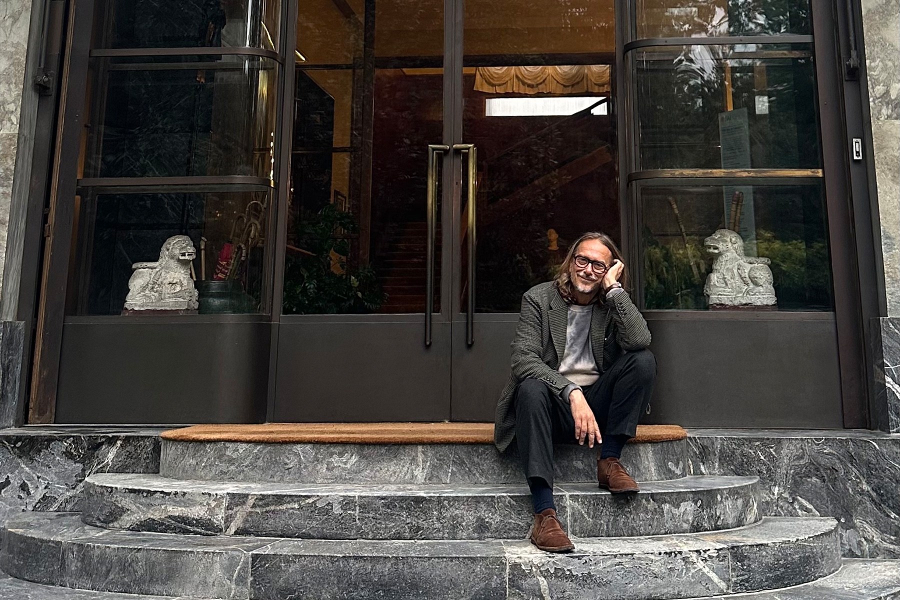 A smiling man with glasses and long hair sits on marble steps in front of a glass door with lion statues.