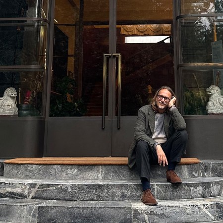A smiling man with glasses and long hair sits on marble steps in front of a glass door with lion statues.