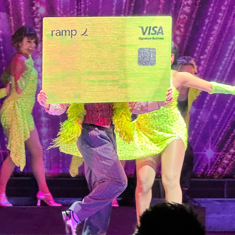 Performers in bright neon outfits on a stage, holding up a large yellow Ramp Visa card.