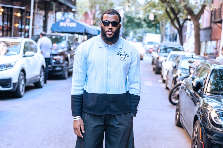 A bearded man in sunglasses and a light blue and black color-block shirt with a panda logo stands on a city street.