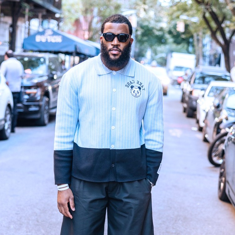 A bearded man in sunglasses and a light blue and black color-block shirt with a panda logo stands on a city street.