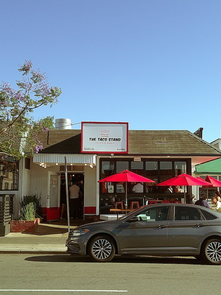The Taco Stand restaurant with red umbrellas and a gray car parked in front on a sunny day.