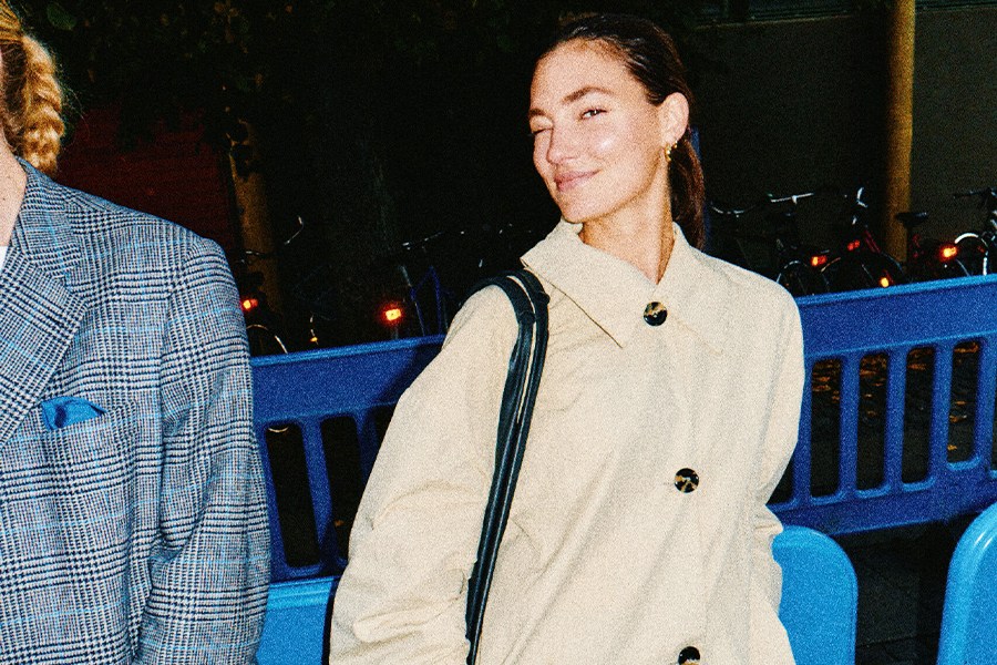 Woman in a beige coat winks, standing next to a person in a blue plaid jacket, with bicycles in the dark background.