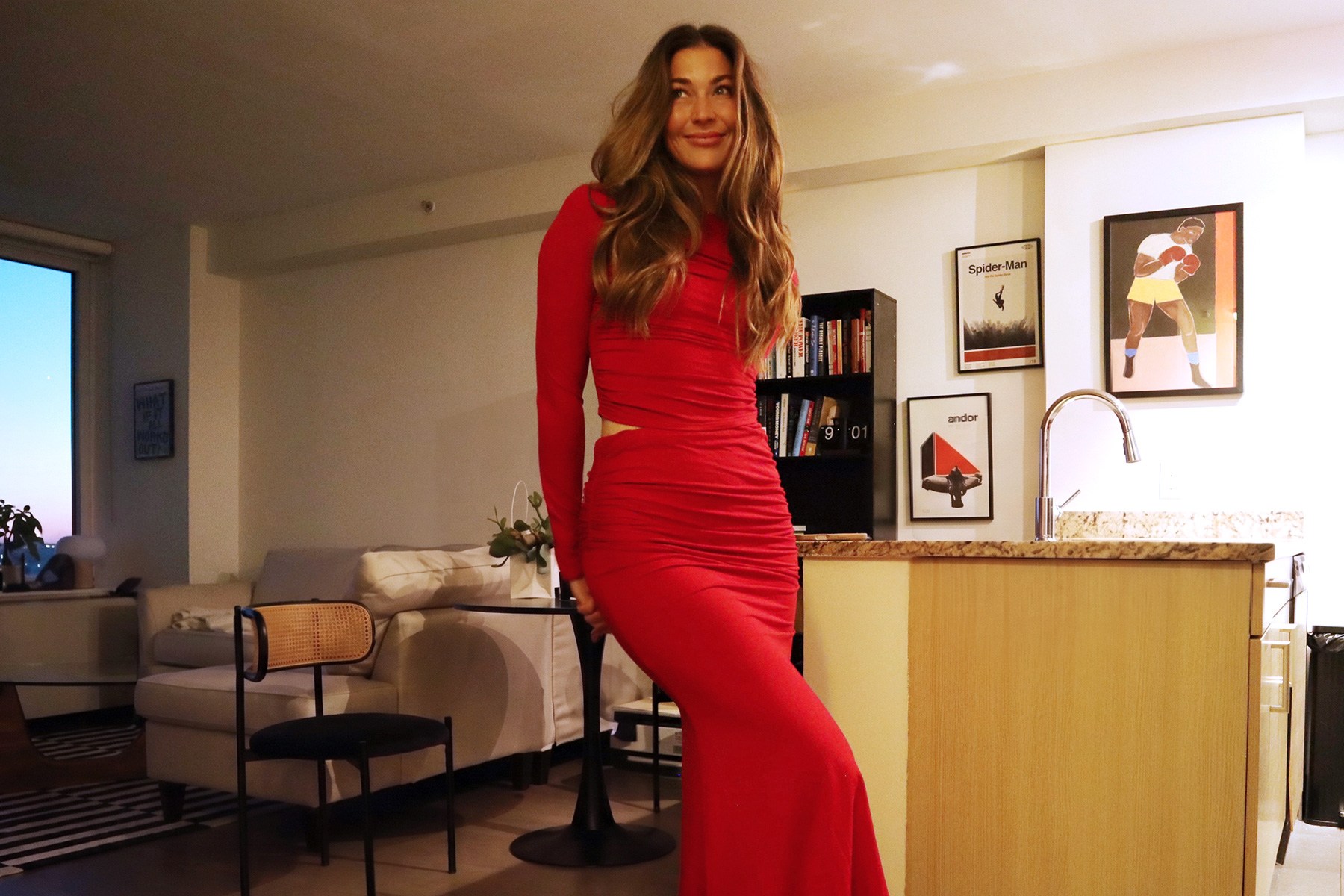 Smiling woman in a long-sleeved red ruched dress with a cutout at the waist, standing in a modern apartment.