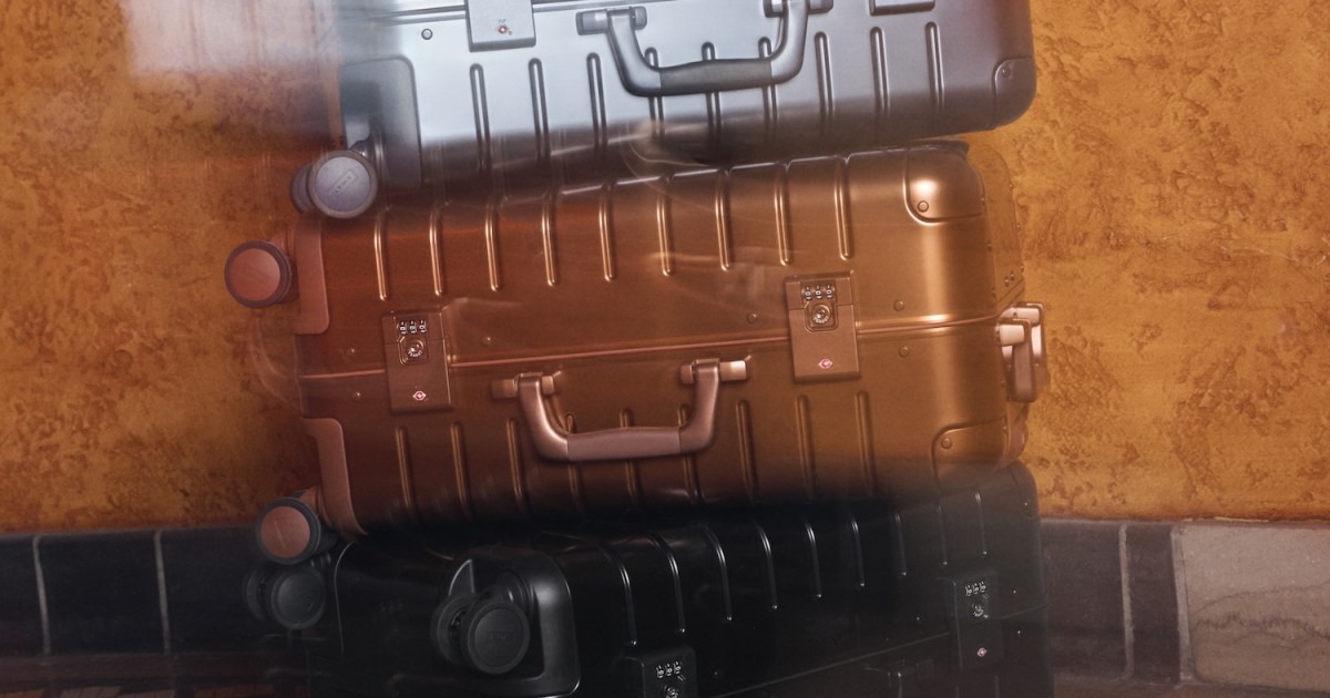 Three Away aluminum suitcases, stacked: silver, bronze, and black, against a textured orange wall.