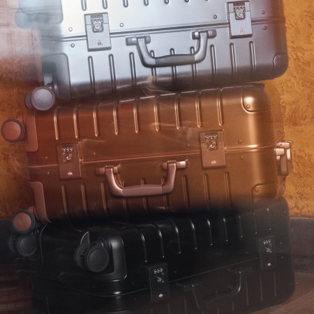 Three Away aluminum suitcases, stacked: silver, bronze, and black, against a textured orange wall.