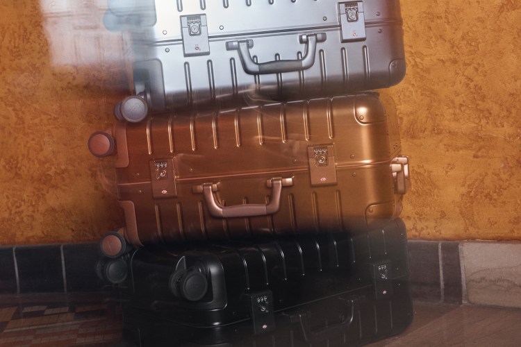 Three Away aluminum suitcases, stacked: silver, bronze, and black, against a textured orange wall.