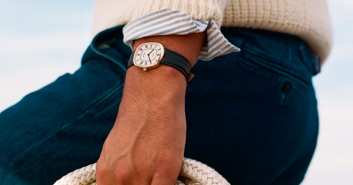 A person wearing a gold watch with a dark green strap, a cream sweater, and blue pants holds a coiled rope.