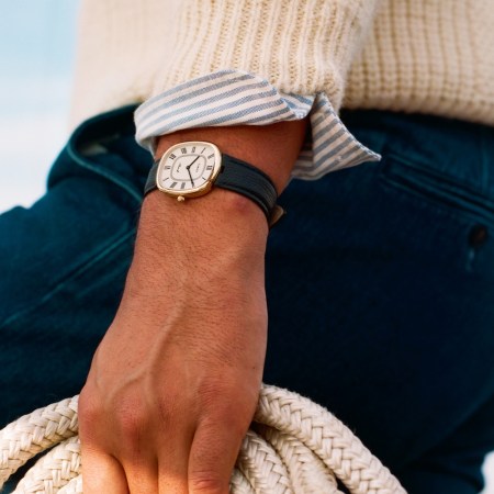 A person wearing a gold watch with a dark green strap, a cream sweater, and blue pants holds a coiled rope.