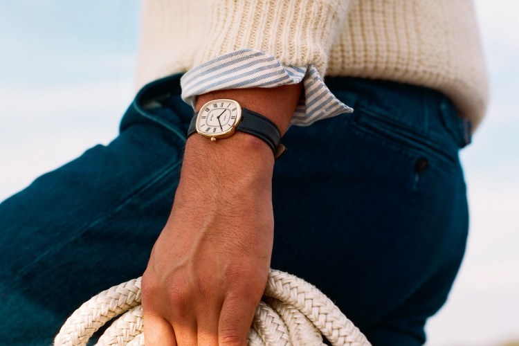 A person wearing a gold watch with a dark green strap, a cream sweater, and blue pants holds a coiled rope.