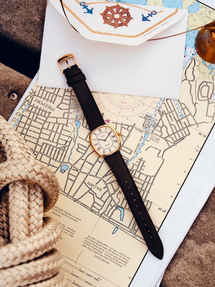 A gold Timex watch with a black leather strap lies on a nautical chart, next to coiled rope and an envelope.
