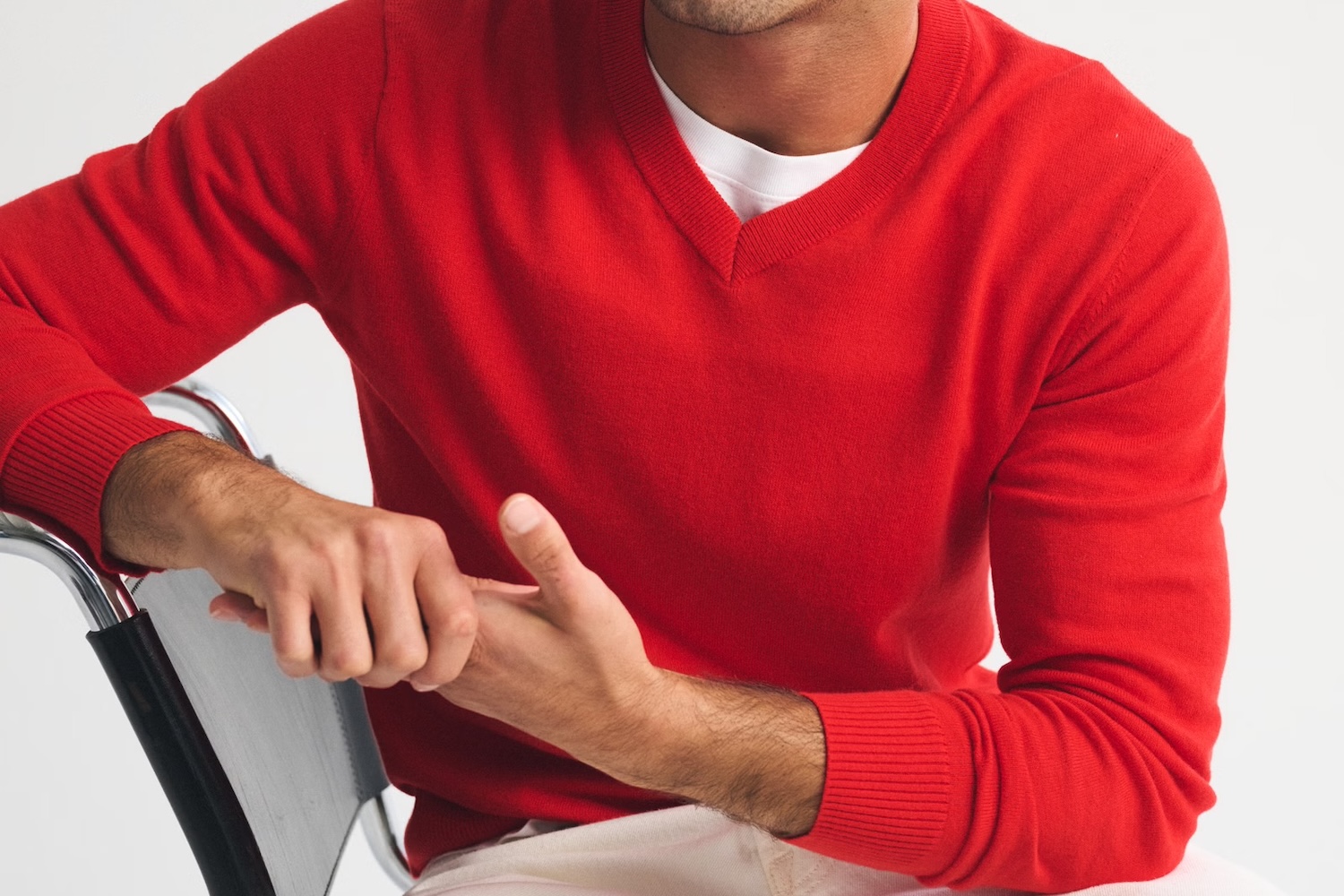 A man in a red V-neck sweater and white undershirt sits with his hands clasped on a chair arm.