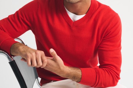 A man in a red V-neck sweater and white undershirt sits with his hands clasped on a chair arm.