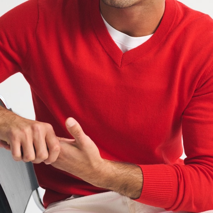 A man in a red V-neck sweater and white undershirt sits with his hands clasped on a chair arm.