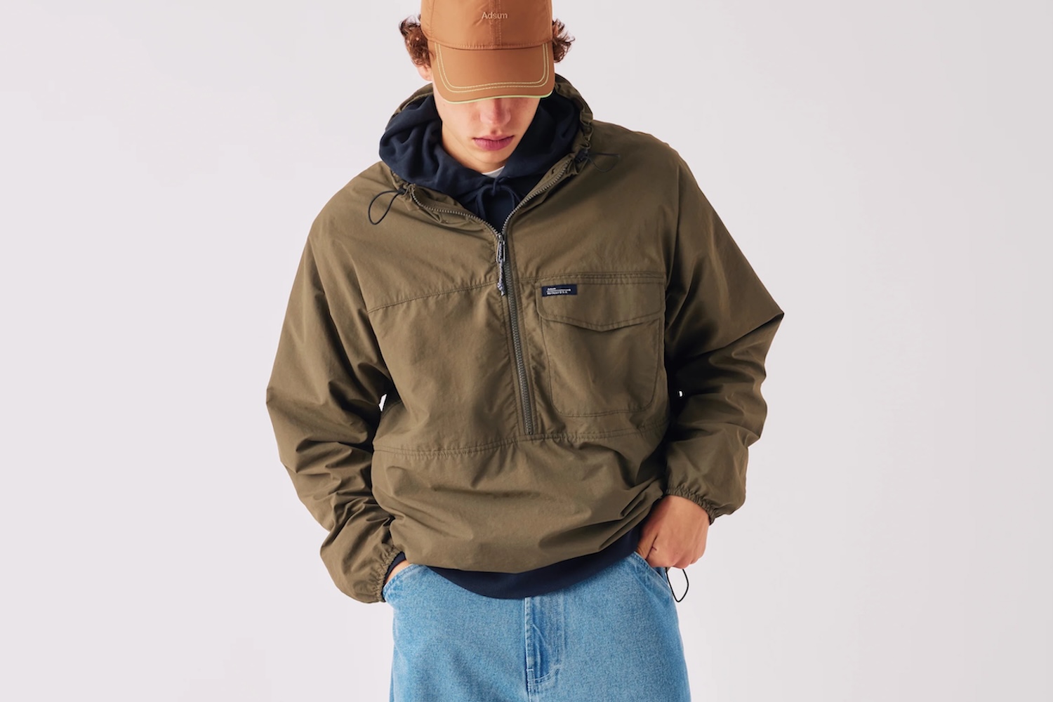 A person in a brown cap, navy hoodie, olive green anorak, and light blue jeans.