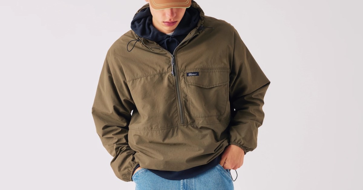 A person in a brown cap, navy hoodie, olive green anorak, and light blue jeans.