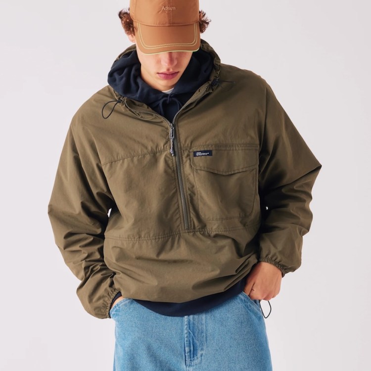 A person in a brown cap, navy hoodie, olive green anorak, and light blue jeans.
