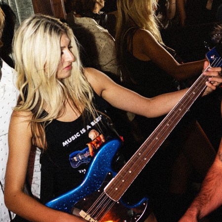 A blonde woman in a "Music is a Blessing" tank top plays a blue glitter bass guitar, flanked by a smiling DJ and a man in a beret.