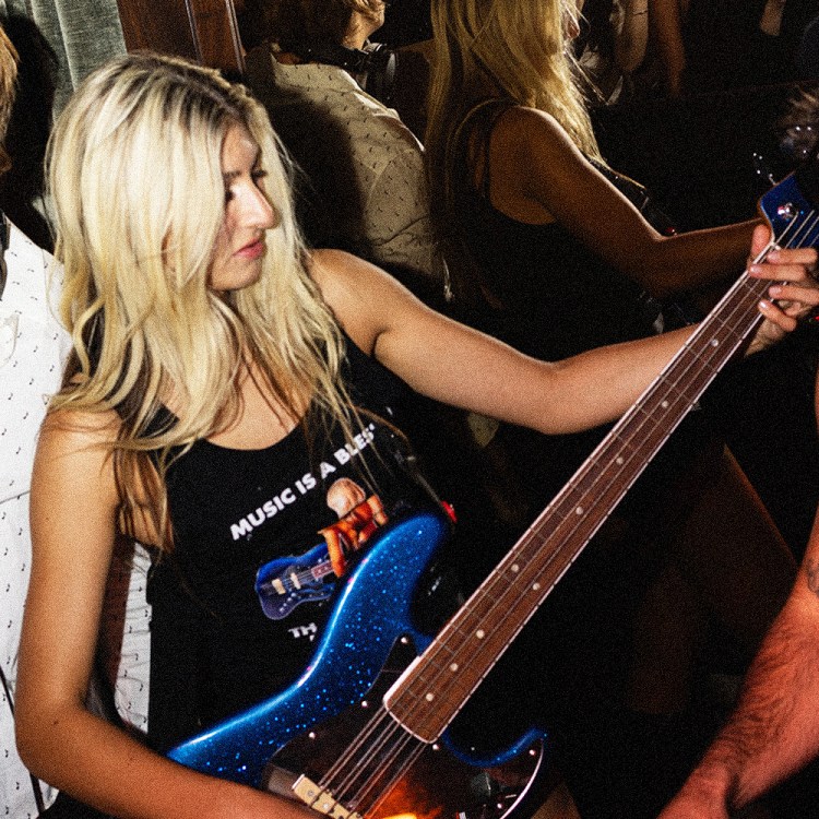 A blonde woman in a "Music is a Blessing" tank top plays a blue glitter bass guitar, flanked by a smiling DJ and a man in a beret.