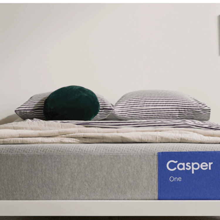 A Casper One mattress with a blue label, striped pillows, and a dark green round pillow on a white bed frame.