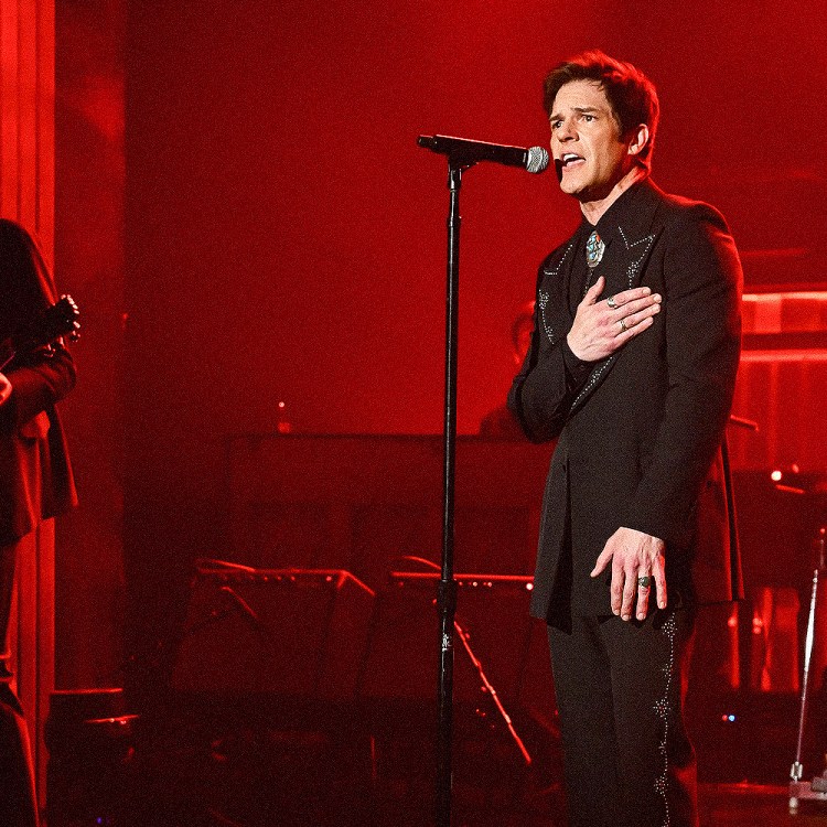 Singer Brandon Flowers in a black suit with a bolo tie and rings, hand on his chest, singing into a microphone on a red-lit stage