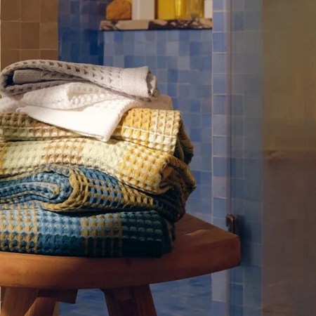 A stack of waffle-weave towels in white, gray, yellow, and blue on a wooden stool in a tiled bathroom.