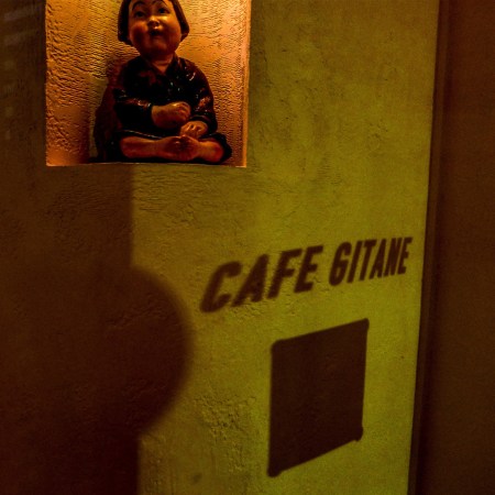 A small statue of a child sits in a niche above the shadow of "CAFE GITANE" on a textured wall.