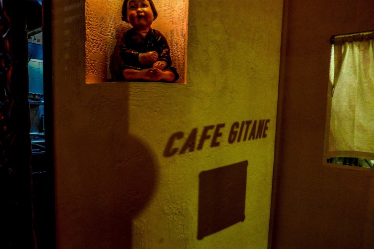 A small statue of a child sits in a niche above the shadow of "CAFE GITANE" on a textured wall.