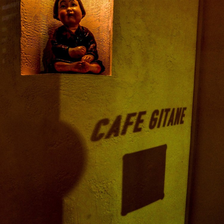 A small statue of a child sits in a niche above the shadow of "CAFE GITANE" on a textured wall.