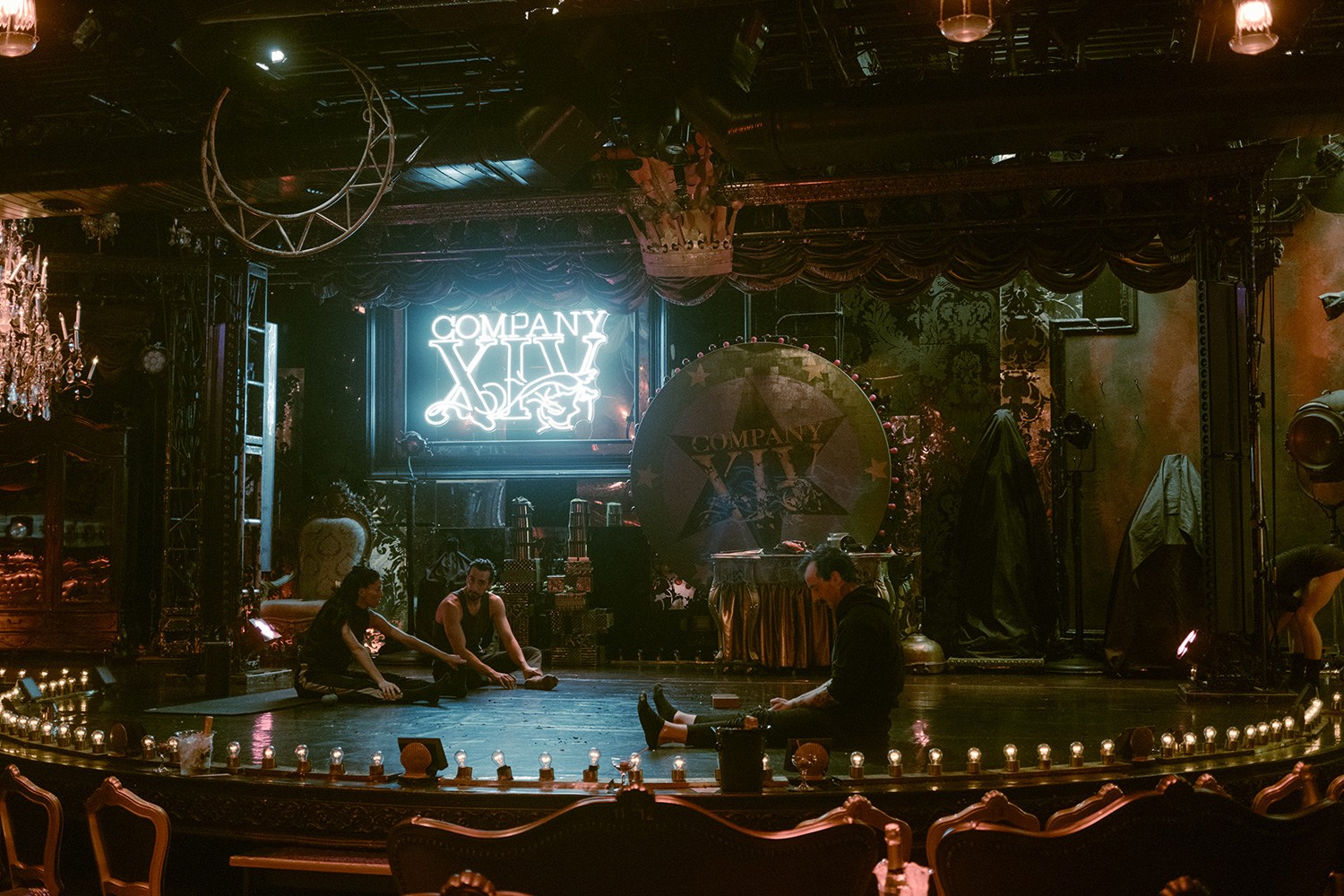Performers stretching on a dimly lit stage with a neon "COMPANY XIV" sign and a large wheel.