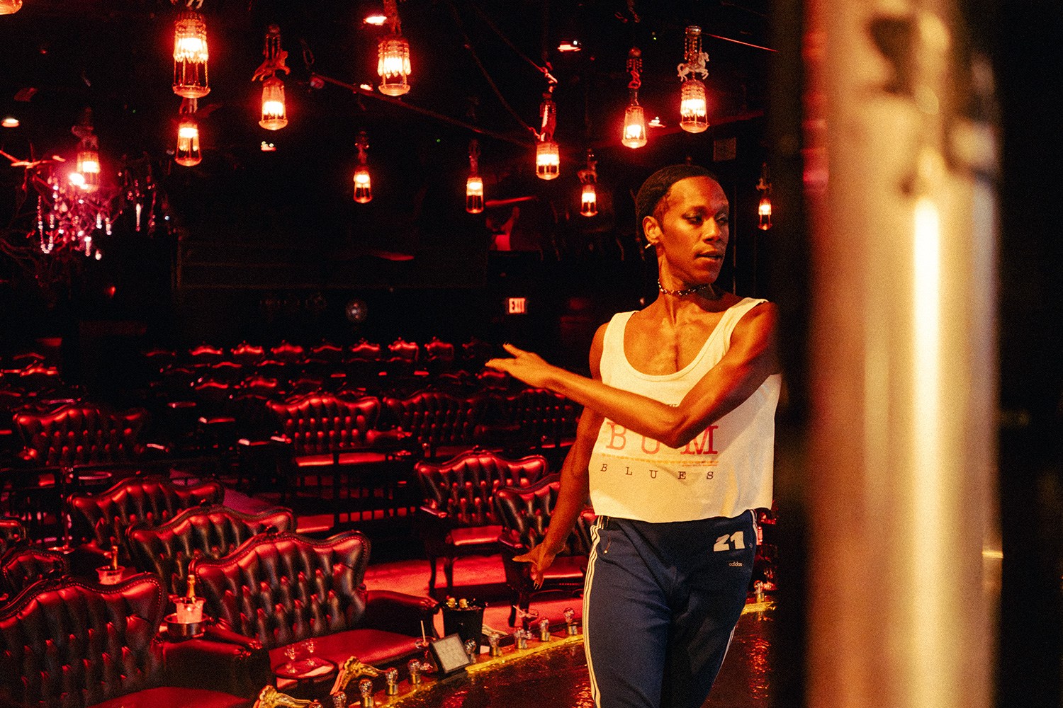 A person in a white tank top and dark pants stands in a dimly lit room with red leather chairs and hanging lights.
