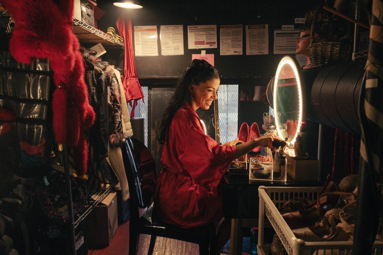 a brunette woman in a red robe smiling and doing her makeup at a vanity