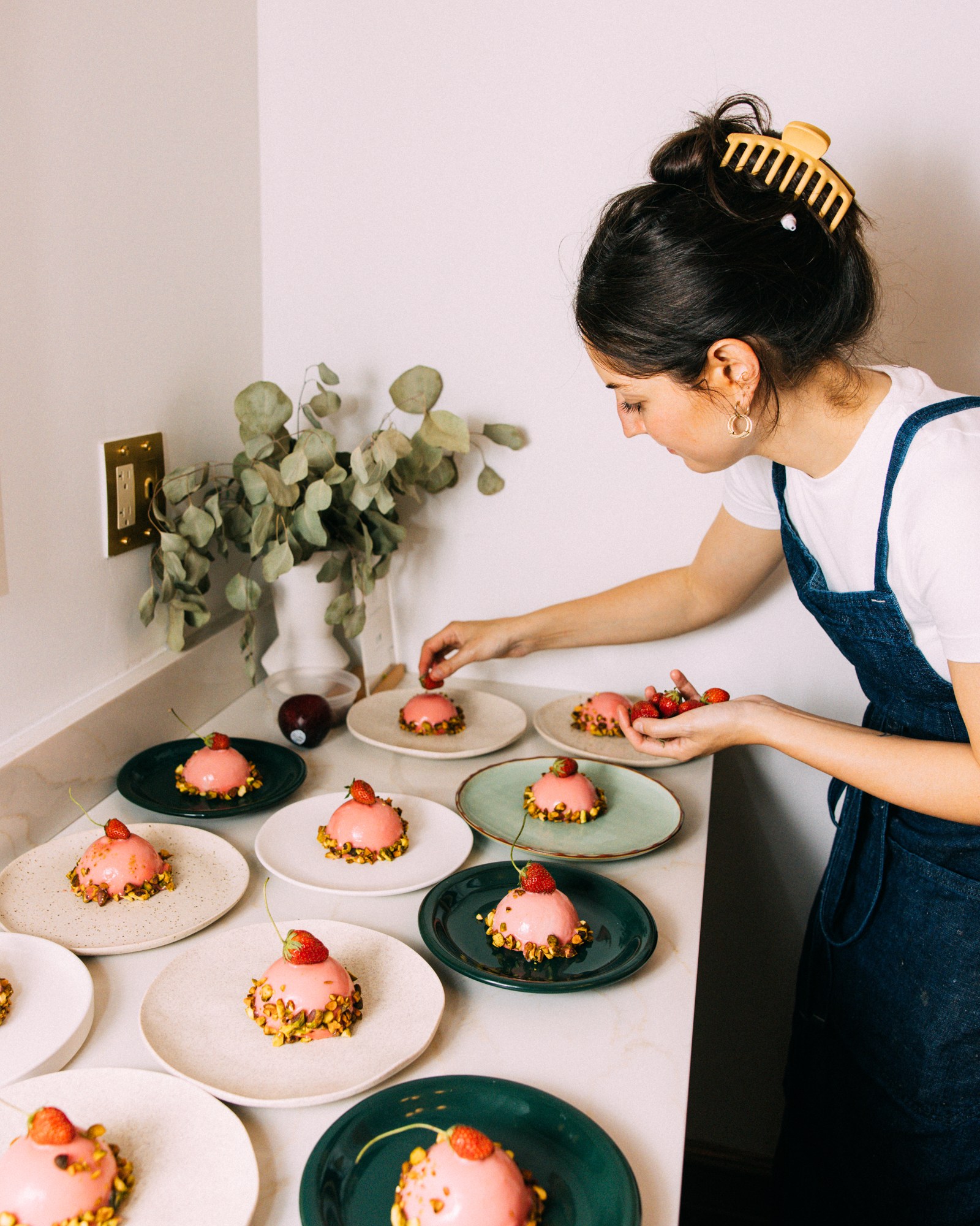 Kendra Vaculin in an apron garnishes several pink, dome-shaped desserts with strawberries and pistachios on a white counter.
