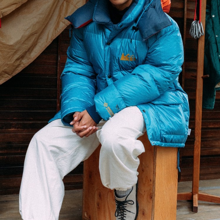 Person in blue puffer jacket and white pants sits on a wooden box, with camping gear nearby.