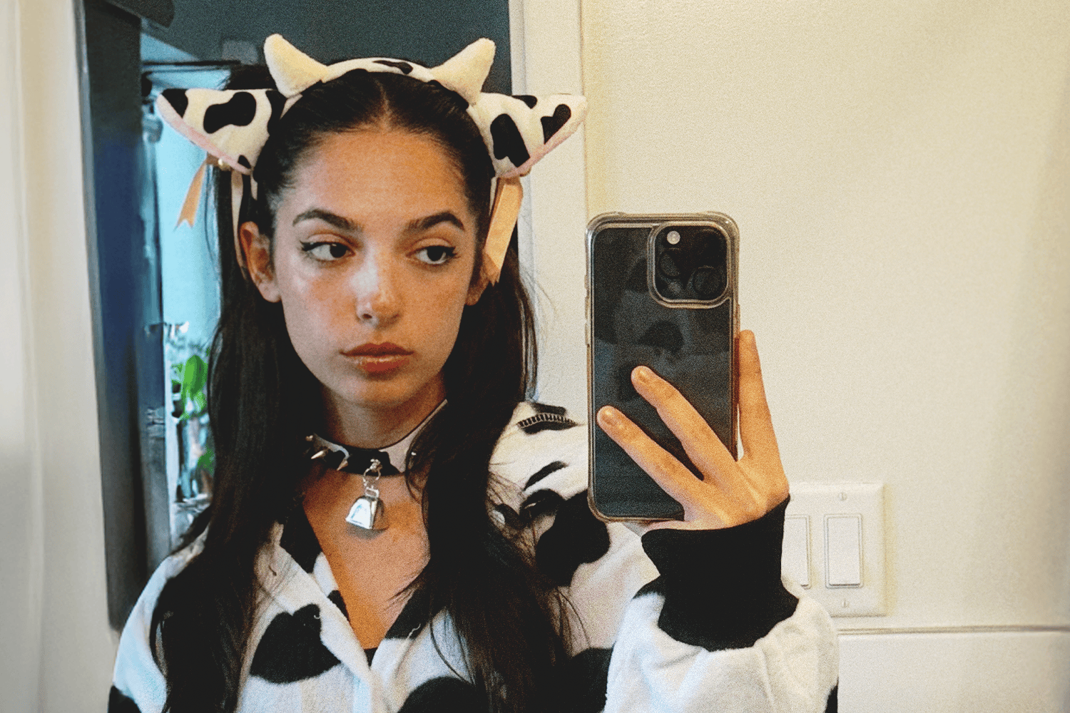 Young woman in a cow costume with a spiked collar and bell takes a mirror selfie, looking away from the camera.