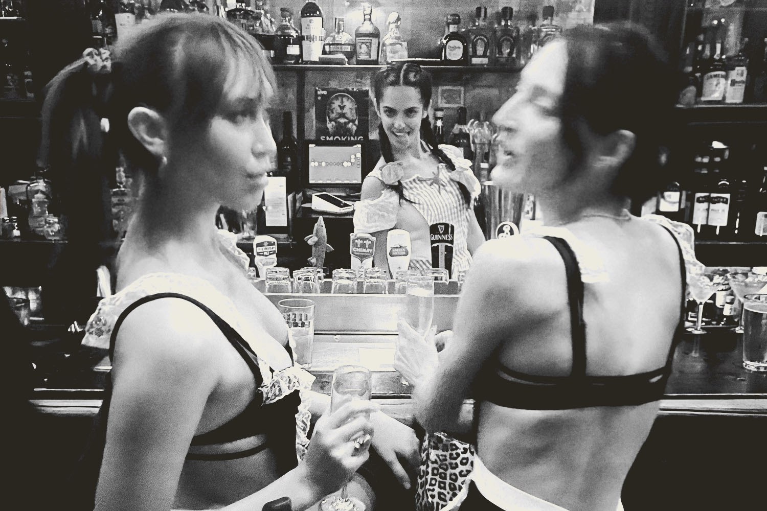 Three women in a bar, two in foreground wearing lingerie, one bartender smiling in background.