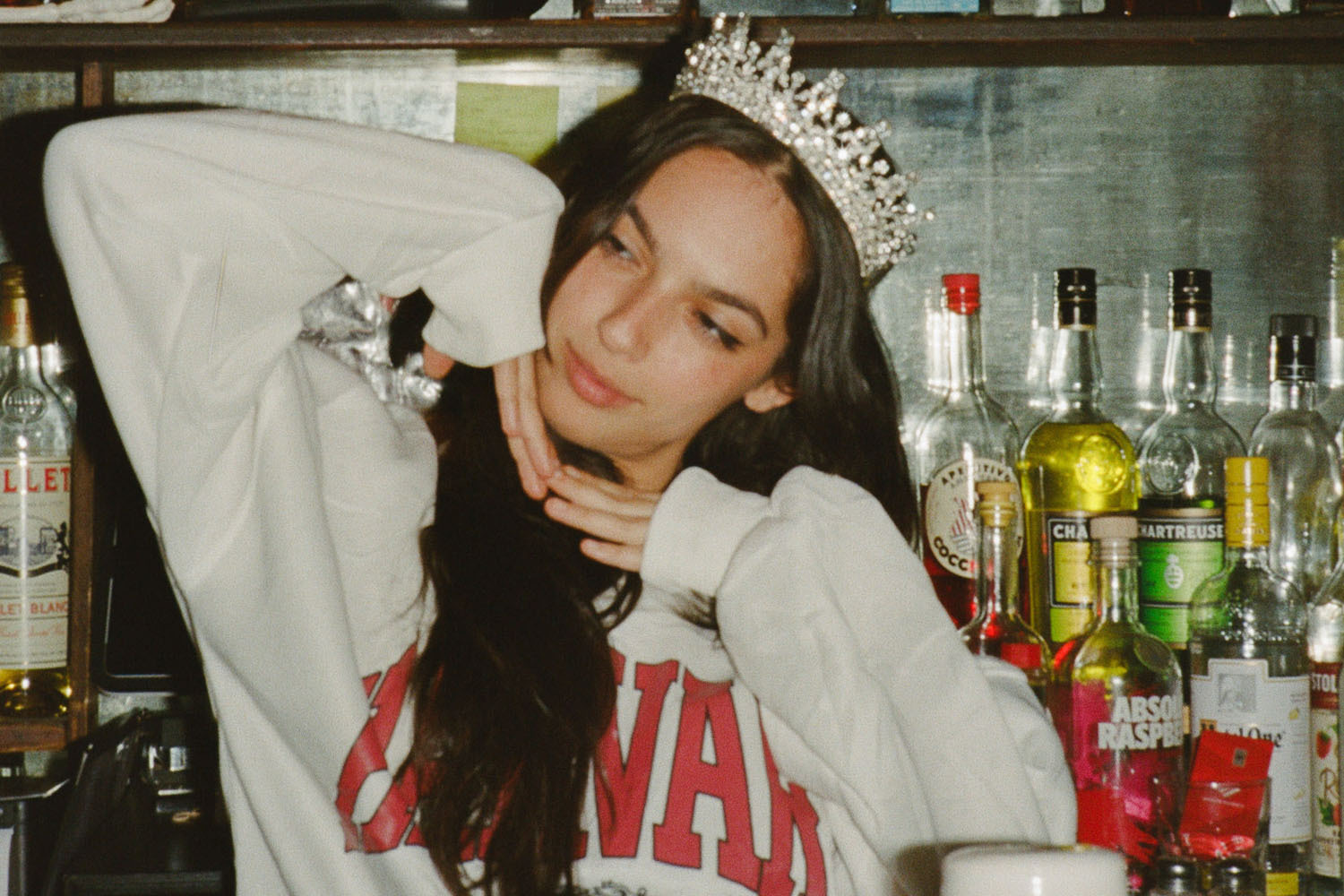 Young woman in a crown and white sweatshirt poses in front of a bar with bottles of liquor.