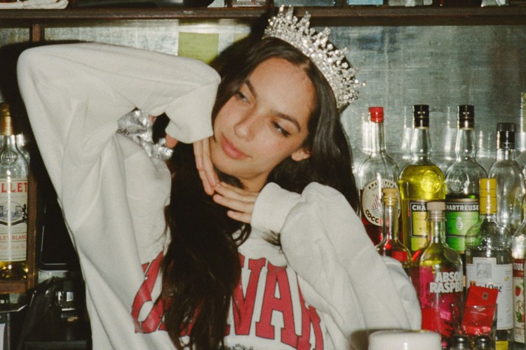Young woman in a crown and white sweatshirt poses in front of a bar with bottles of liquor.