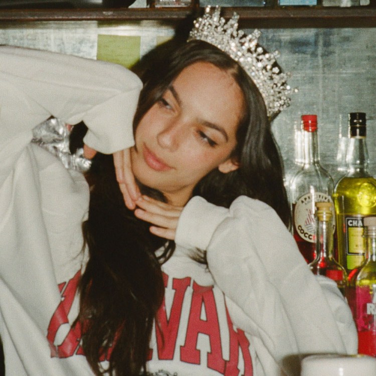Young woman in a crown and white sweatshirt poses in front of a bar with bottles of liquor.
