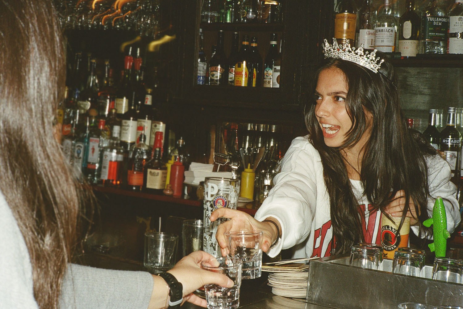 A woman in a tiara smiles while handing a shot glass to another person across a bar.